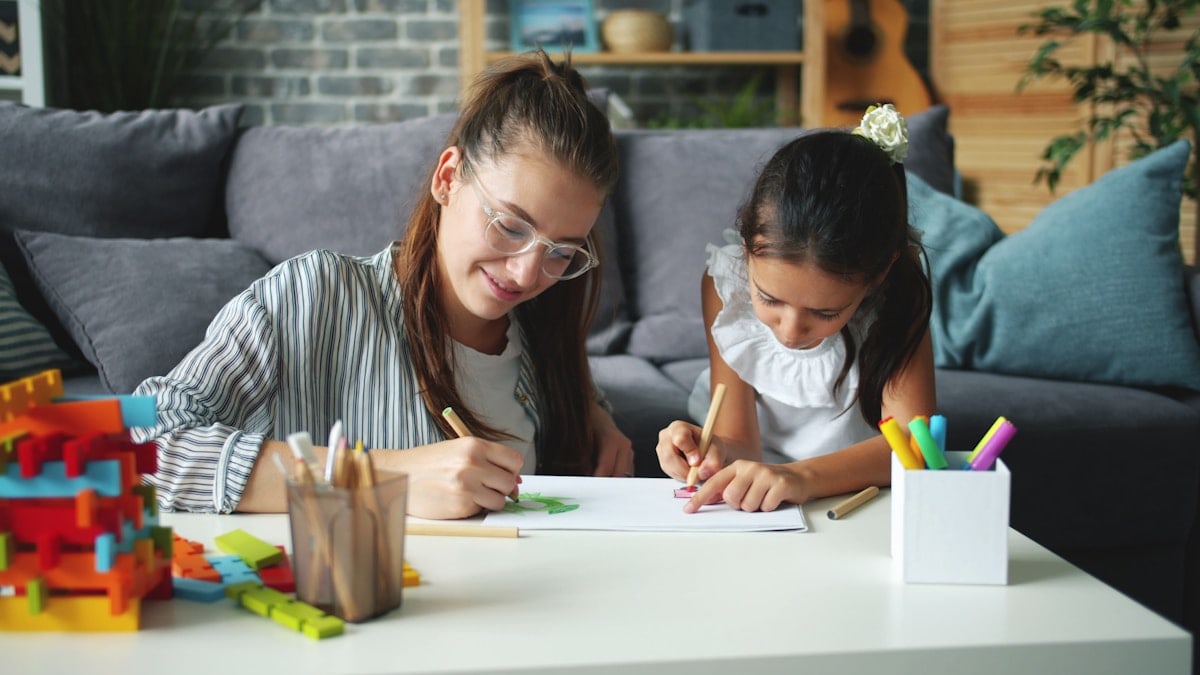Cute little girl creative child is drawing picture while caring mother is helping expressing love. Leisure time at home, people and happy family concept.